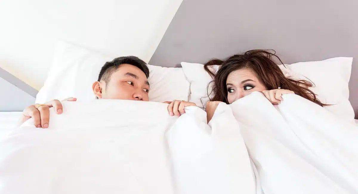 Two anonymous hands intertwined on a bed beside a small vibrator and a bedside lamp
