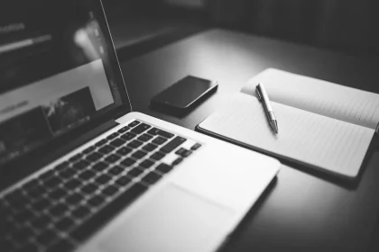 Hands typing on a laptop at a newsroom desk with a smartphone and notepad